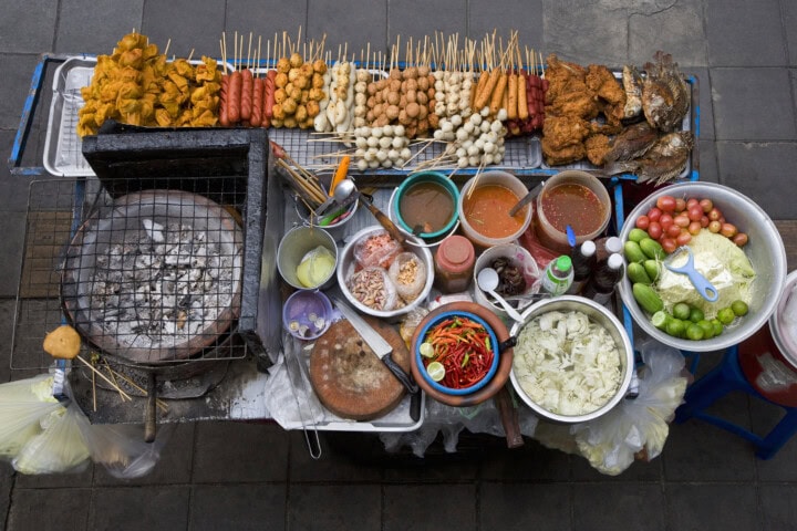 A bustling street food stall reminiscent of Vietnam, with an assortment of skewers, condiments, and fresh vegetables displayed on a table, along with a grill sizzling on the left side.
