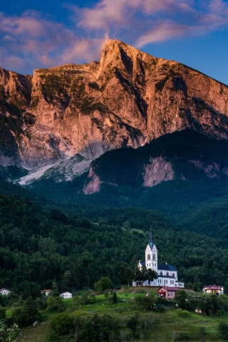 A tall church with a pointed steeple stands amid a forested landscape in Slovenia, with a large, rocky mountain illuminated at sunset in the background.