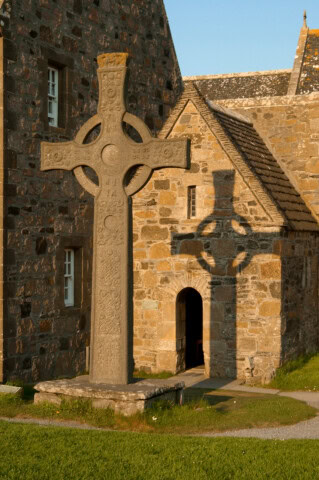 A large stone Celtic cross is positioned outside a historic stone building in Scotland, casting a shadow on the wall. The building has an arched doorway and multiple small windows. The surroundings include a grassy area.