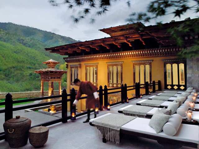 A person arranges lanterns on a terrace with lounging chairs, set against the backdrop of an ornate yellow building and lush green mountains, capturing the essence of Bhutan's unique charm and inviting tourism.