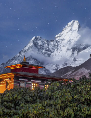 A traditional building with illuminated windows sits in front of a snow-capped mountain under Nepal's starry sky.