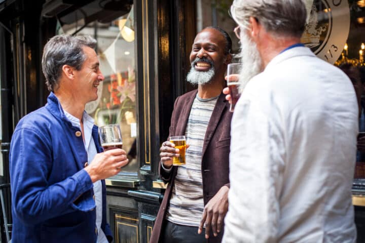 Three men stand outside an Irish pub, each holding a glass of beer, and engage in a lively conversation.