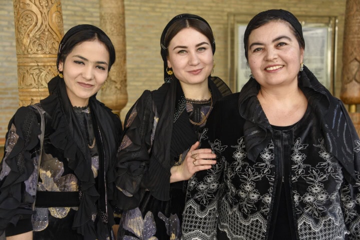 Three women wearing traditional Uzbek clothing stand side by side, smiling at the camera. Ornate wooden columns are visible in the background.