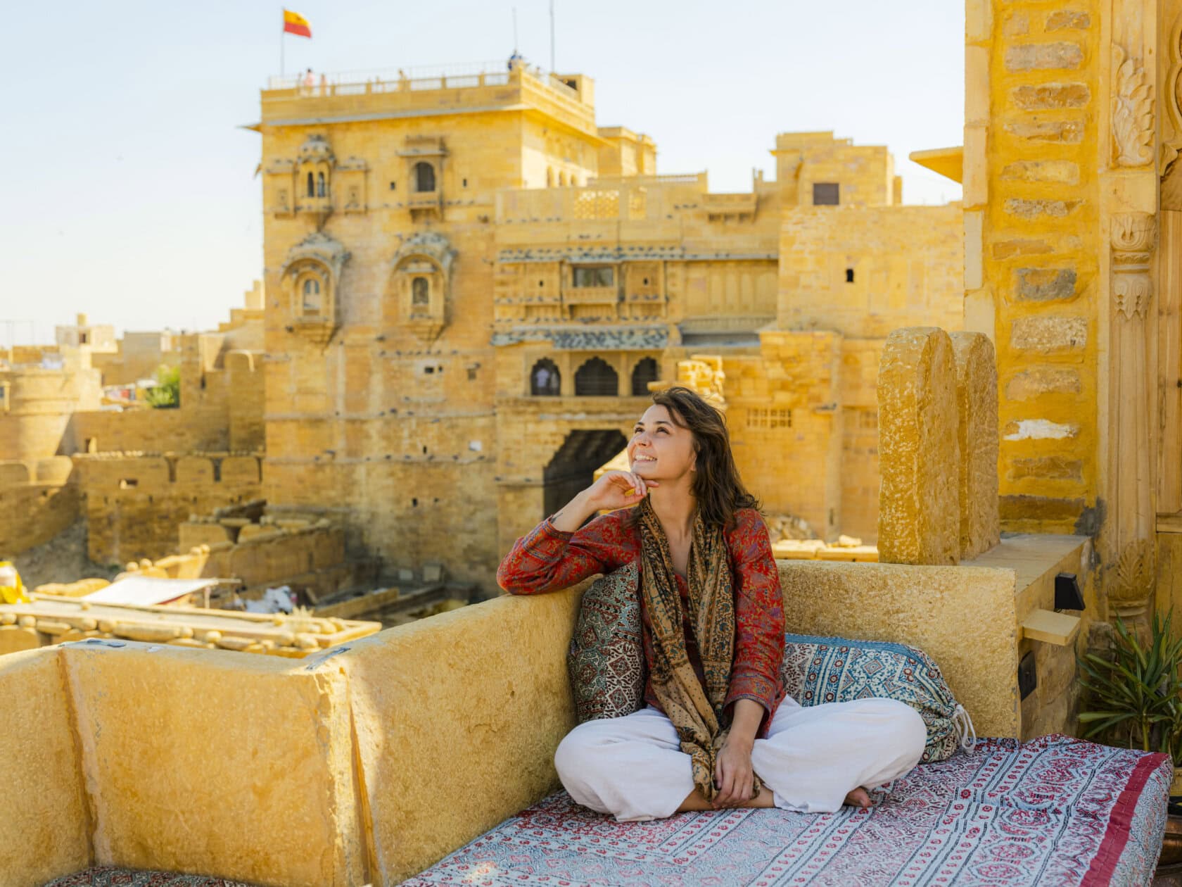 A person sitting cross-legged on a cushioned terrace in Rajasthan, looking up thoughtfully. Historic sandstone buildings rise majestically in the background under a clear sky, making this moment feel like it belongs in a beginner's guide to India.