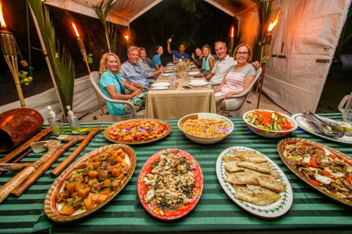 A group of people sit around a long table inside a large tent, with an array of food dishes laid out on a striped tablecloth in the foreground. The atmosphere buzzes with excitement as they celebrate their Palauan feast together.