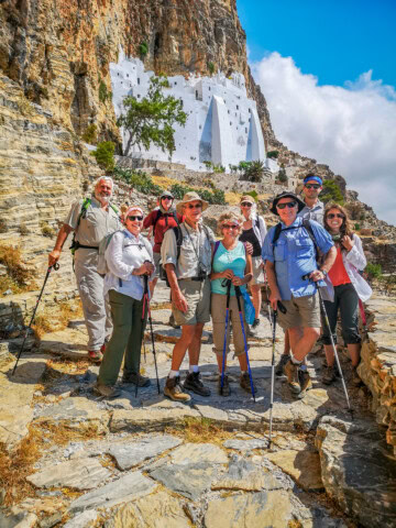 A group of hikers standing on a rocky path in front of a white building built into a cliff in Greece, posing for a photo with hiking poles and outdoor gear.