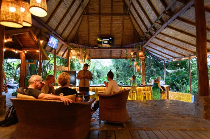 People relax and socialize in a thatched-roof lounge with wicker furniture, surrounded by lush Costa Rican greenery. There are hanging lights, a bar in the background, and a TV on the wall.