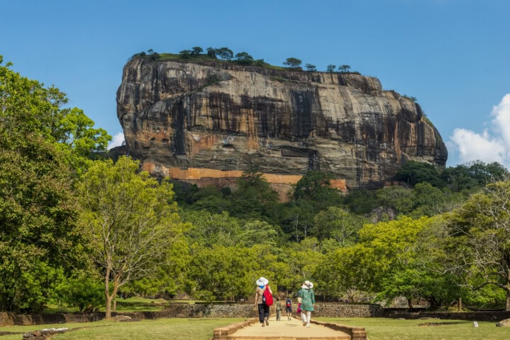 Three people walk on a path towards a large rock formation surrounded by trees at Sigiriya, Sri Lanka, on a clear day.
