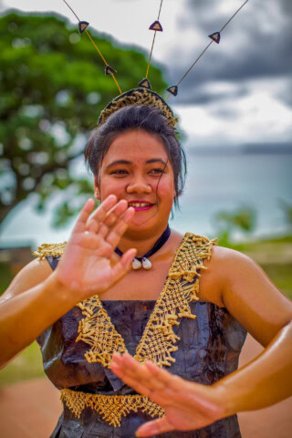 A person wearing traditional attire and headgear performs a dance with a scenic background featuring a tree and the ocean, reminiscent of the vibrant culture found in the Polynesian islands.