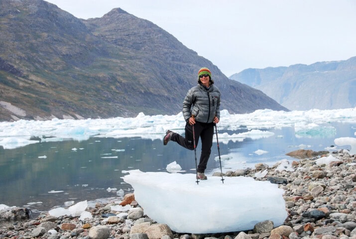 A traveler balances on one leg atop a small ice formation by a rocky shore, surrounded by the stunning landscape of icy waters and mountains in Greenland. Perfect for an adventurous tourism snapshot!