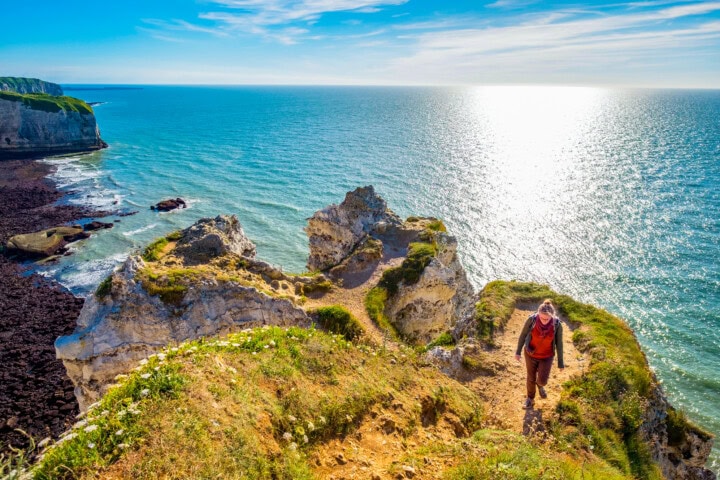 A person wearing an orange and black outfit hikes along a cliffside trail overlooking a sunlit ocean on a clear day, enjoying one of France's stunning travel destinations.