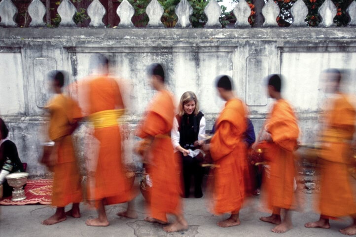A row of young monks in orange robes walk past a seated person against a stone wall in Laos, creating a motion blur effect—a captivating travel moment that embodies the essence of local tourism.