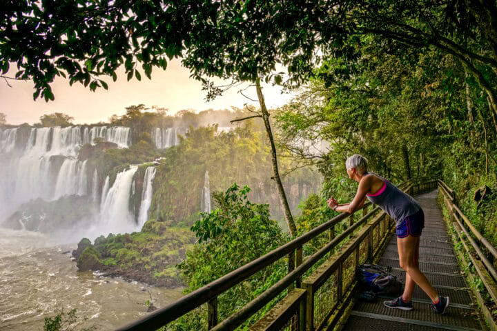 A person in athletic wear leans on a railing, capturing a photo of Argentina's majestic waterfalls amidst lush greenery from a metal walkway.