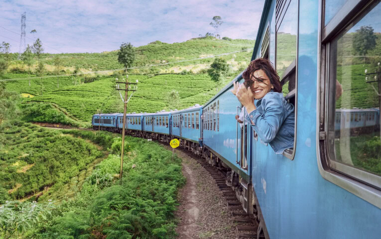 A person leans out of a blue train window, smiling and waving while traveling through the lush green hills of Sri Lanka, showcasing the charm of travel and tourism in the region.