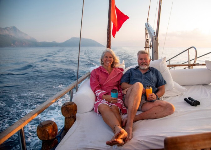 Two people relaxing on the deck of a boat at sea, sitting on a white mattress with Turkey's mountainous coastline in the background, holding cups and smiling.