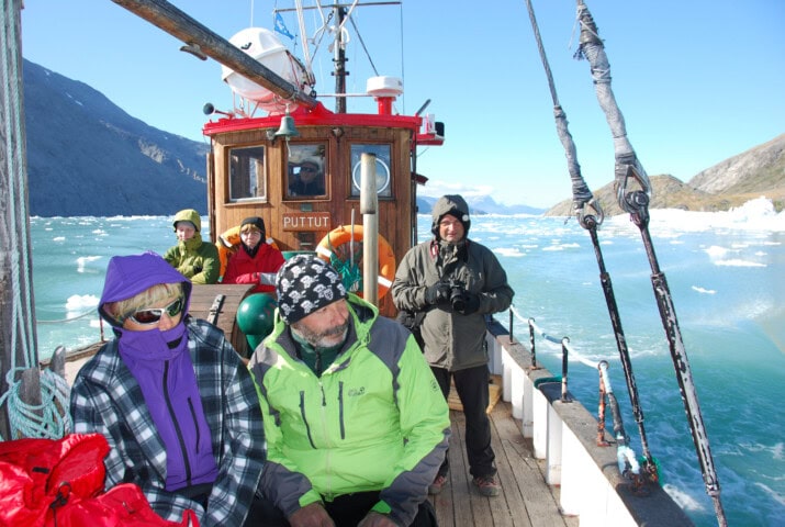 People are on a small boat named 'Piittut' in the icy waters of Greenland. Most are wearing winter jackets and hats. Icebergs and mountainous terrain are visible in the background.