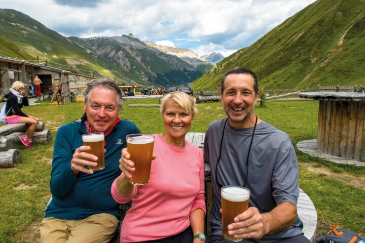 Three people sitting outdoors in the picturesque mountains of Slovenia, smiling and raising glasses of beer. They are seated on wooden benches with rolling hills and lush greenery in the background.