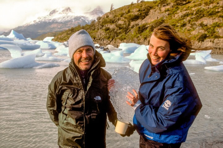 Two people in outdoor gear smile while standing by floating ice chunks in Chile, with mountainous terrain in the background. One person holds a piece of ice.
