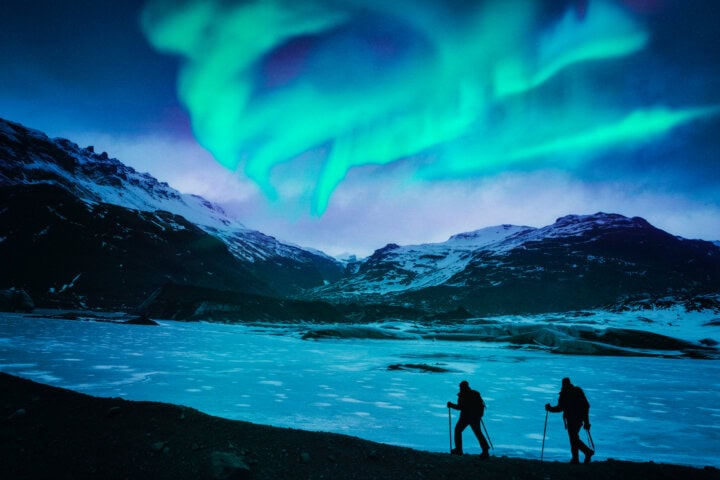 Two individuals hiking with poles on a snowy landscape under a vivid display of green northern lights in the sky. The mountainous terrain in the background highlights the breathtaking beauty of Iceland, making it a prime destination for winter tourism and travel enthusiasts.