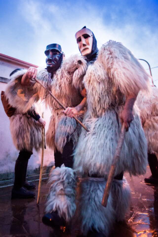 People in fur costumes and masks holding staffs during a traditional celebration at dusk in Italy.