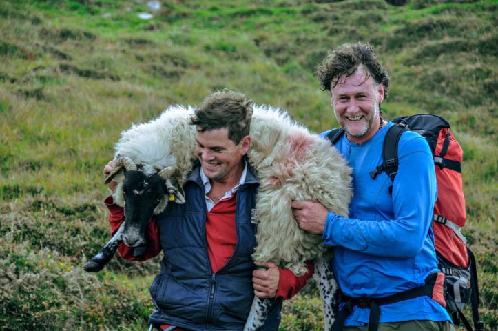 Two men stand outdoors, one carrying a sheep on his shoulders. They are smiling, dressed in outdoor attire, with a lush grassy field typical of Ireland behind them. This charming scene is perfect for anyone interested in travel and tourism in the Emerald Isle.