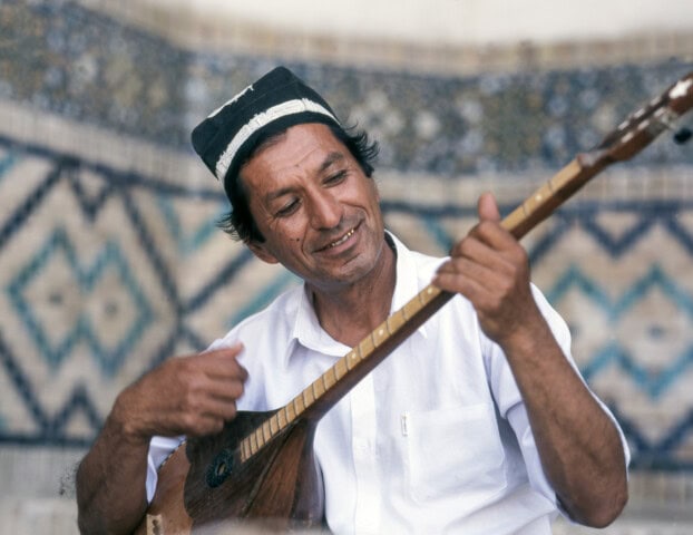 A man wearing a hat and a white shirt plays a stringed instrument outdoors in Uzbekistan, with the background featuring beautiful patterned tiles.