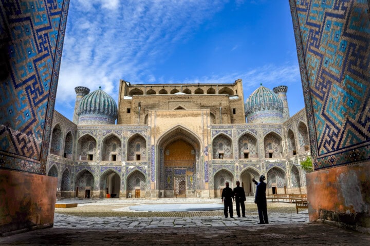 View of an ornate historical building with blue domes and intricate tile work, framed by archways. Three people stand in the foreground, silhouetted against the structure. This breathtaking scene captures the essence of Uzbekistan and beckons travelers to explore Central Asia's rich heritage.