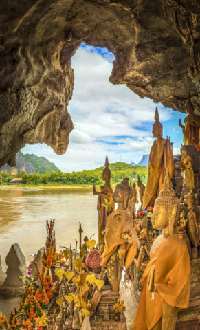 Buddha statues inside a cave with offerings, overlooking a river and landscape through the cave opening.