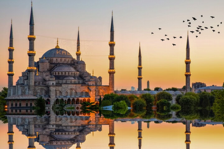 The image shows the Blue Mosque in Istanbul during sunset, with its six minarets silhouetted against the sky and a flock of birds flying nearby, reflected in a body of water in the foreground—a perfect snapshot for travel enthusiasts exploring Turkey's marvels.
