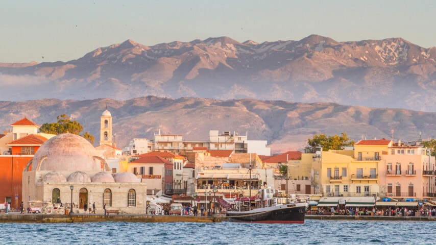 A coastal town in Greece with colorful buildings, a domed structure, and a mountainous backdrop at sunset. Several boats and people are visible near the water.