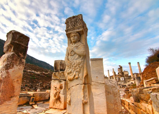 Ancient ruins in Turkey featuring a tall, detailed stone statue amid scattered columns and stonework, with a partly cloudy sky in the background.
