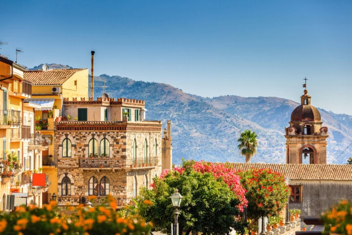 A scenic view of an Italian town with colorful buildings, a bell tower, blooming flowers, palm trees, and mountains in the background under a clear blue sky.