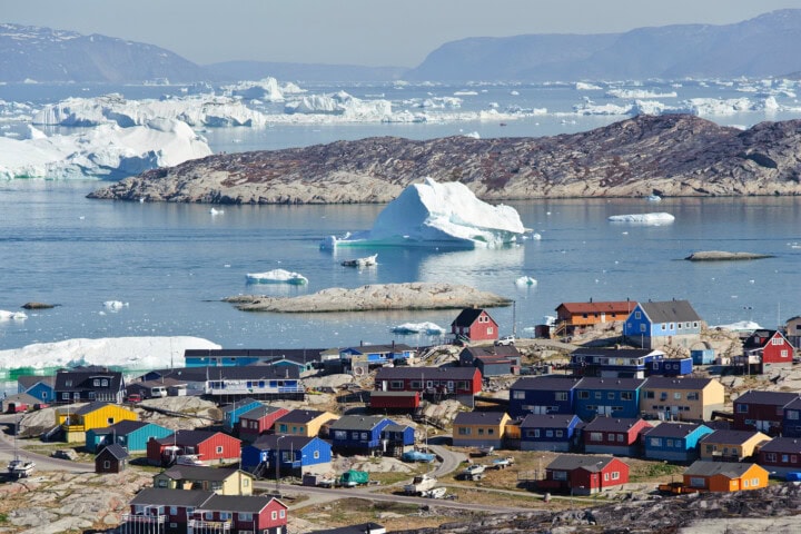 A coastal town in Greenland with brightly colored houses sits next to icy waters, where large icebergs float nearby. Rugged, rocky terrain and distant mountains are visible in the background.