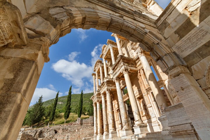 The ancient Library of Celsus in Ephesus, Turkey, viewed through a stone archway, with a clear blue sky and green hills in the background.
