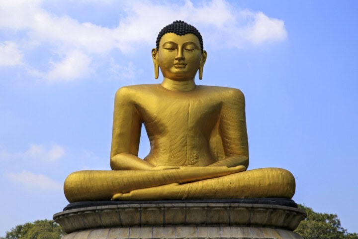 A large golden Buddha statue, seated in a cross-legged position, graces Sri Lanka's landscape against a backdrop of blue sky with scattered clouds. The statue rests majestically on a stone pedestal.
