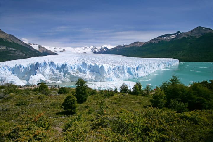 A vast glacier in Argentina spans from left to right, bordered by a turquoise lake and green forest, extending to mountain peaks under a cloudy sky in the background.