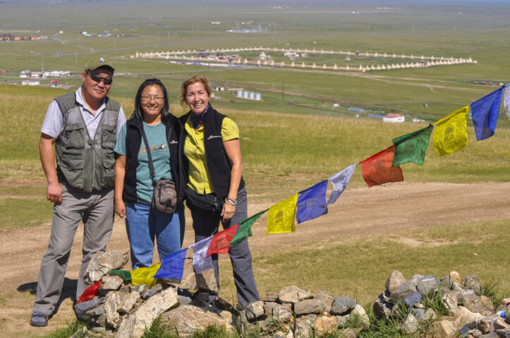 Three people stand together in front of a rocky mound adorned with colorful prayer flags, capturing the essence of travel in Mongolia. A large, open plain with traditional structures is visible in the background under a clear sky, offering a glimpse into the region's rich tourism potential.