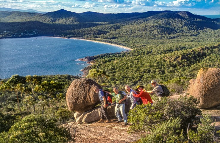 A group of people stands on a rocky hill in Tasmania, posing together with a large boulder. A scenic view of a beach, ocean, and forested hills is visible in the background.
