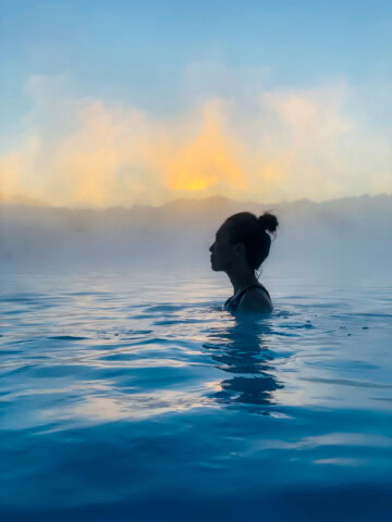 Person with hair in a bun standing in a large body of water with fog and a bright sky in the background, epitomizing the serene beauty of Iceland. This picturesque scene is perfect for anyone's travel or tourism dreams.