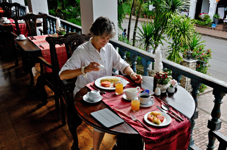 A person enjoys breakfast on a patio in Laos, featuring toast, fruit, juice, and coffee. The setting boasts wooden flooring, red tablecloths, and lush greenery in the background.