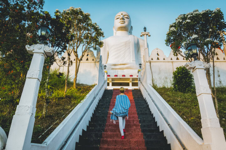 A person walks up the steps toward a large white Buddha statue, framed by trees and a white fence, against a clear Sri Lankan sky.
