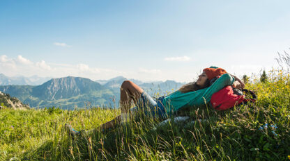 Person lying on a grassy hill with a scenic mountain view in the background, wearing a green sweater and an orange hat, with a red backpack by their side, embracing secrets of healthy living.