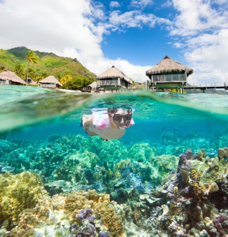 A person wearing snorkeling gear swims in the clear turquoise waters of French Polynesia above a coral reef near stilted huts and lush green mountains, with a partly cloudy sky above.