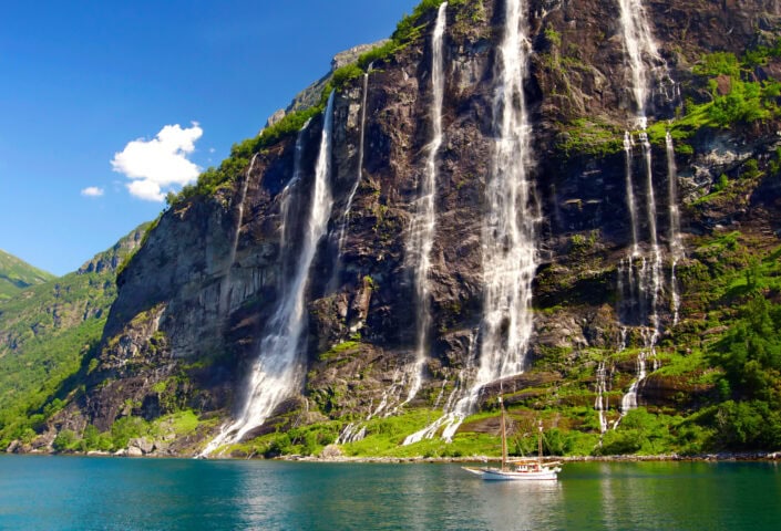 A white sailboat floats on calm blue water near tall streams of waterfalls cascading down a steep, rocky, green mountainside under a sunny sky.