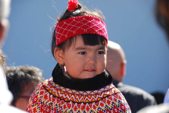 A young child with a red patterned headband and a colorful embroidered outfit stands out against the blue background, surrounded by blurred onlookers, evoking the vibrant spirit of Greenland.